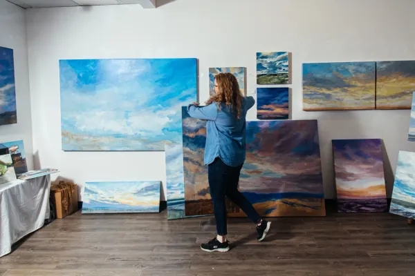 Artist arranging large landscape paintings in a studio gallery space, surrounded by vivid skies and cloudscapes displayed on easels and walls.