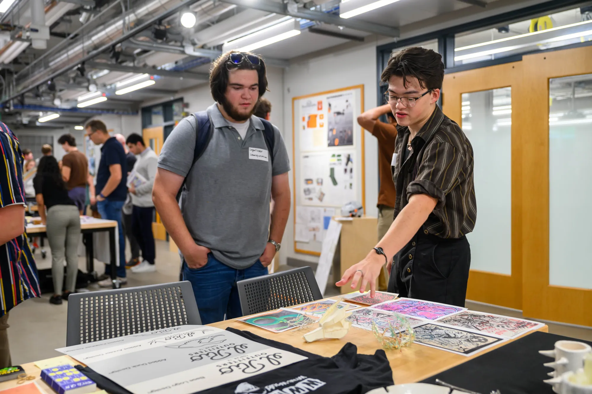 A student explains their printmaking and illustration work to a visitor at a showcase event. Prints, t-shirts, and other creative pieces are displayed on a table in front of them. Other attendees interact in the background of the studio space.