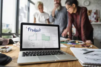 Open laptop displaying product sketches alongside the word Product. Designers are working together in the background behind the laptop