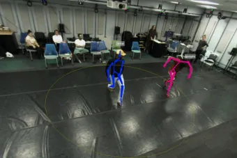 Two dancers perform on a black mat in a motion capture studio, with their movements tracked by colored digital outlines—blue for one dancer labeled “Sinclair” and pink for the other labeled “Mustapha.” Several people observe from chairs in the background, and tracking equipment is visible on the ceiling and walls.