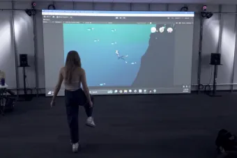A person with long hair stands in front of a large projection screen displaying an underwater-themed video game. The game shows a diver swimming near a coral reef with fish surrounding them. The person mimics a swimming motion, suggesting interactive gameplay.