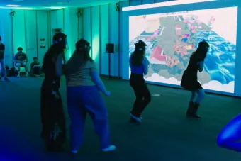 Four students wearing motion-tracking caps interact with a large projected video game that features a colorful landscape with butterflies, houses, and waves. The room is bathed in green and blue lighting, with several observers seated along the walls.