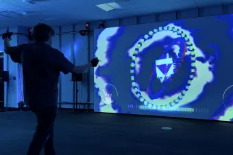 A student wearing motion-tracking gloves stands in front of a large screen, interacting with a colorful, abstract digital interface featuring 3D geometric shapes and swirling patterns. The space is illuminated in blue lighting, with audio equipment positioned around the room.