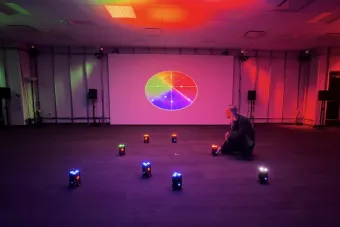A student crouches in a dark room illuminated by red and purple lighting, surrounded by small LED light modules arranged in a circle. A color wheel is projected on a large screen in the background, suggesting the interaction of light, color, and spatial audio in an immersive installation.