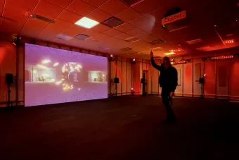 A person stands in a red-lit room holding a wand-like controller, interacting with a large projection of a virtual stone-walled chamber. Glowing particles spiral toward a circular portal on screen, suggesting motion-based interaction with a magical or fantasy-themed digital environment.