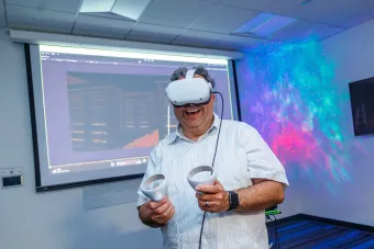 A person smiles while wearing a VR headset and holding motion controllers in a classroom. Behind them, a projected screen displays a virtual environment, and colorful light patterns illuminate the wall.
