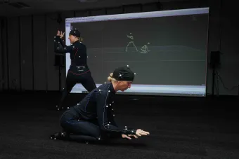Two performers in motion-capture suits demonstrate choreography in a dark studio, with their movements digitally rendered in real time on a large screen behind them. The space highlights immersive technology and interdisciplinary collaboration.