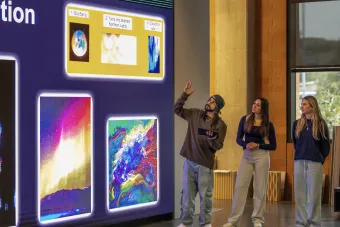 A student presenter gestures toward a large digital display while two other students look on, with the screen showing a three-step visual sequence labeled "Big Bang," "Turns into abstract Northern Lights," and "Clouds to rain" alongside colorful abstract artwork panels.