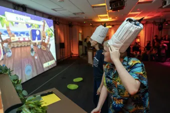Two participants wearing tall chef hats rigged with motion sensors play a virtual cooking game projected on a large screen showing a countdown timer and animated kitchen scene.