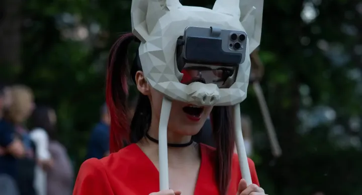 A person wearing a futuristic, geometric bat mask with a smartphone attached to the front, acting as a digital visor. The mask is white with angular facets, covering the entire head. The person, dressed in a striking red outfit, holds two white rods extending from the mask. Their mouth is open in an expressive pose. The background is blurred with an outdoor setting and a crowd of people, suggesting a live event or performance.