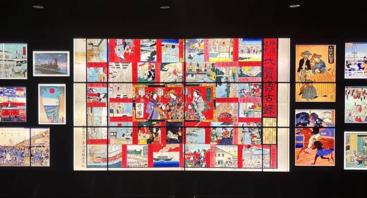 A large digital display wall showcasing a vibrant collage of Japanese woodblock prints. The central composition features a colorful, multi-panel artwork filled with scenes of traditional life, people in elaborate attire, and architectural elements, framed by red borders and Japanese text. Surrounding the center panel are smaller prints, depicting trains, Western visitors, marketplaces, parades, and scenic landscapes. The background is black, emphasizing the illuminated art pieces, which highlight a cultural