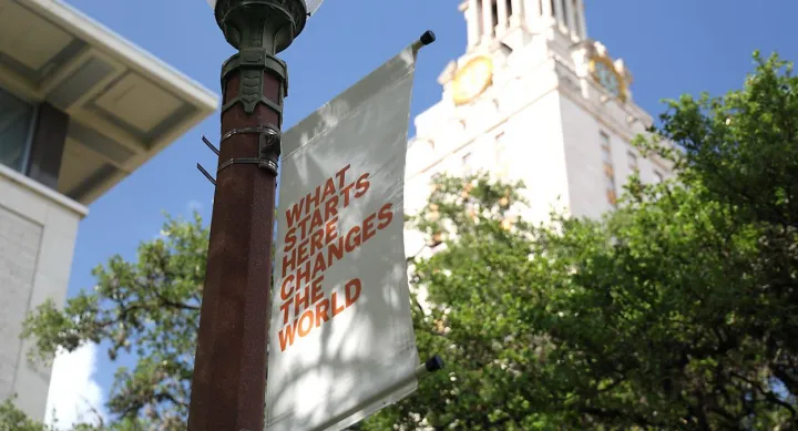Banner on a UT Austin campus lamp post reads “What starts here changes the world” in bold orange text, with the university tower and green trees in the background under a clear blue sky.